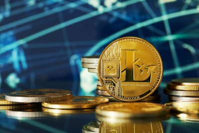 A close-up, eye-level shot of a shiny gold Litecoin coin positioned centrally in the foreground. The coin features a large 'L' symbol, a mining pickaxe icon, and the word 'LITECOIN'. Several other gold and silver-colored coins are stacked and scattered around it, reflecting on the polished dark surface. The background is a blurred, dark blue with illuminated, abstract geometric lines, creating a digital and futuristic ambiance.