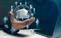 A hand gently cradles a glowing blue wireframe globe, upon which a white 3D model of a classical bank building is prominently placed. The globe and its surroundings are adorned with multiple bright blue upward-pointing arrows and white percentage symbols, conveying a strong sense of growth and increasing financial metrics. The background is a dark, blurry blue, with a portion of a laptop visible on the right side.