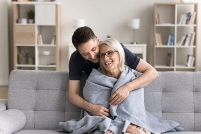 Let me take care of you. Caring attentive grown up millennial man son wrapping shoulders of grateful mom sit resting on sofa with warm woolen blanket protect loved parent from cold at cool living room