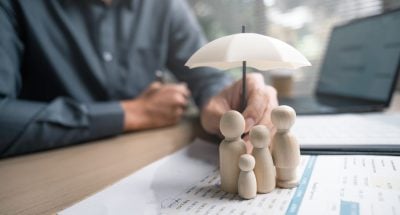 Family insurance plan protect. Help medicine protection. Father mother children insurance. A man is holding a white umbrella over a family of three wooden figures.