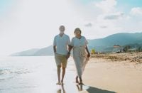Happy senior couple enjoying their vacation, walking hand in hand along the ocean shoreline at sunset, reflecting love, companionship, and retirement joy