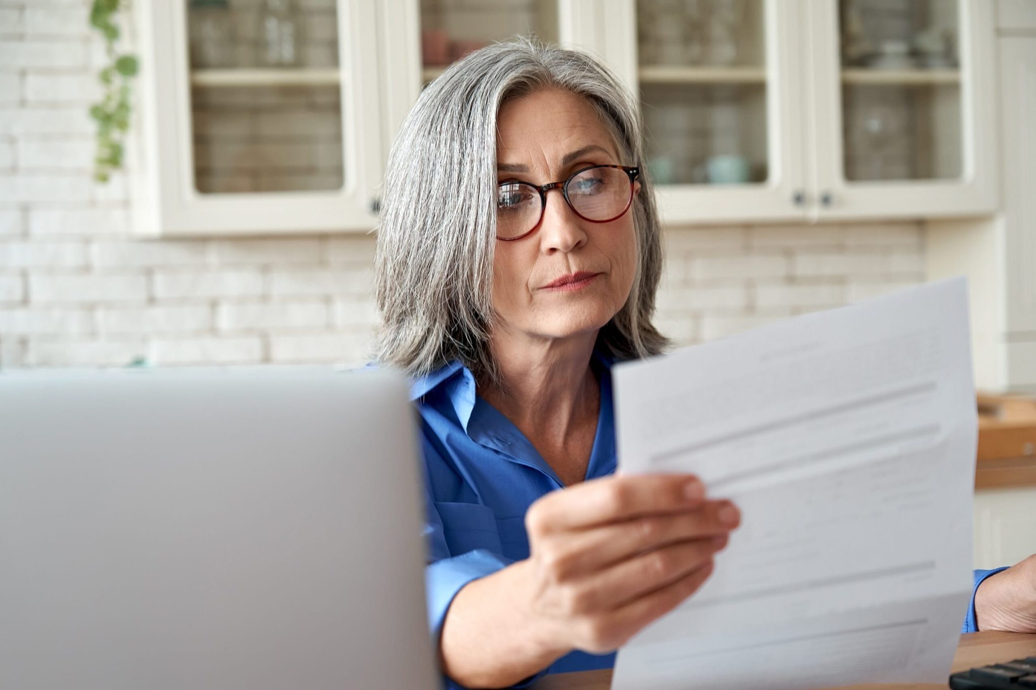 Serious worried mature woman reading paper bill pay online at home. Old lady holding bank letter managing account finances, calculating taxes, planning loan deb