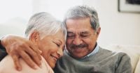 Hug, care and senior couple in house, love and smile with retirement, happiness and bonding together. Apartment, embrace and mature woman with elderly man on sofa, relax and comfortable in lounge