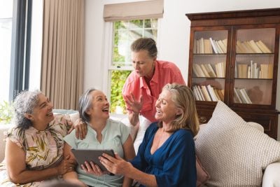 Diverse senior female friends sitting at home, one holding tablet. Sharing light moments and laughter, they enjoy their time together with Caucasian, Asian, and biracial backgrounds, unaltered