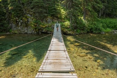 Backpacker Crosses Suspension Bridge Across The Outlet Of Elizabeth Lake In Glacier National Park