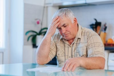 A distressed-looking older man with gray hair sits at a light blue glass table, his left hand resting on his forehead, and his right hand near some white papers. He is wearing a light-colored plaid short-sleeved shirt and appears to be in a kitchen or dining area with blurred white cabinets and a pink orchid in the background.