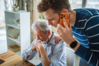 Young man standing and calling emergency services on a cell phone, assisting an elderly man sitting at a table with a pained expression, clutching his chest