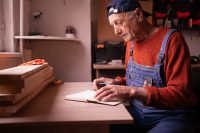 Thoughtful senior wood master writing planning day worker craftsman in workshop carpenter thinking art. copy space