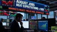 A focused male trader in a suit sits at a desk, looking at multiple computer monitors displaying stock charts and financial data. Above him, a large blue digital ticker displays white text reading 'AUGUST JOBS BEAT EXPECTATIONS: +162K' and red text 'MARKETS REACT: YIELDS RISE, STOCKS SLIP. -1.0%'. Another screen on his desk shows a chart titled '10 YEAR TREASURY YIELD HIT 4.77%'. Other traders are visible in the background, also at desks with monitors, in a dimly lit office environment.