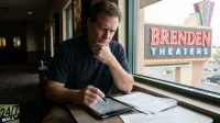 A man in a dark polo shirt sits by a window, looking intently at a tablet and papers. His left hand is on his chin in thought, while his right hand holds a stylus over the tablet. Outside the window, a large sign for 'BRENDEN THEATERS' is visible. The interior is dimly lit, typical of a theater lobby.