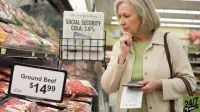 A senior woman with gray hair, wearing a beige jacket and green shirt, stands in a supermarket meat aisle. She holds a calculator and a grocery list in her left hand, and a package of ground beef in her right, looking down thoughtfully. A price sign in the foreground displays 'Ground Beef $14.99'. In the background, a newspaper rack shows a headline 'SOCIAL SECURITY COLA: 3.6%'.