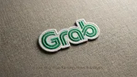 Close-up, slightly elevated shot of an embroidered Grab logo on a textured light brown fabric. The logo is green with a white outline and depicts the word 'Grab' in a stylized, connected script. The fabric background has a visible woven texture.