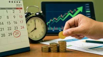 A close-up shot on a wooden desk showing financial elements. A white desk calendar for 2021 has the date '29' circled in red. Beside it, a dark alarm clock shows the time around 8:45. In the foreground, three ascending stacks of gold coins are visible, with a hand placing an additional gold coin on the tallest stack. In the blurred background, a tablet screen displays a green stock market chart with a prominent upward-pointing arrow. Financial documents and a green and gold pen also rest on the desk.