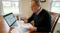 A senior man with glasses and a black sweater sits at a wooden table, holding and reading financial papers. To his left, an open laptop displays an Excel spreadsheet with green, white, and red cells, along with various bar and pie charts. A beige coffee mug, a calculator, and a pen are also on the table. In the background, a large window shows a blurred view of a suburban neighborhood with houses.