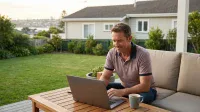 A middle-aged man with short brown hair, wearing a mauve polo shirt and blue jeans, sits smiling faintly while typing on a silver laptop on a wooden slatted outdoor table. A gray coffee mug rests near the laptop. He is seated on a light beige outdoor sofa. In the background, a lush green lawn, various garden plants, a light-colored house, and a blurred cityscape or coastline are visible under a bright sky.