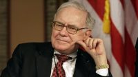 A close-up portrait of Warren Buffett, an older man with light gray hair and glasses, looking to his left with a pensive expression. He is wearing a dark suit, a white shirt, and a red patterned tie. His right hand is resting on his cheek, and he has a gold watch on his left wrist. In the blurred background, a red and white striped American flag with a yellow tassel is visible.