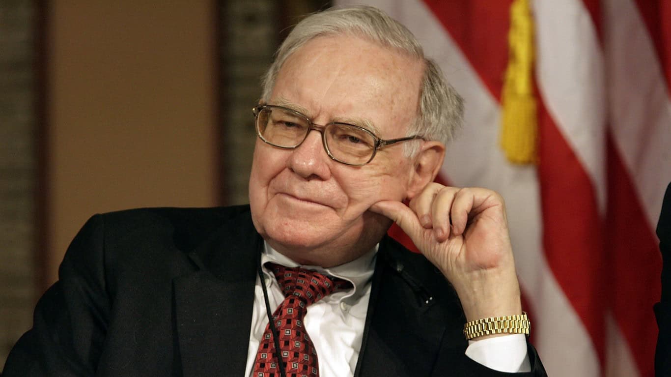 A close-up portrait of Warren Buffett, an older man with light gray hair and glasses, looking to his left with a pensive expression. He is wearing a dark suit, a white shirt, and a red patterned tie. His right hand is resting on his cheek, and he has a gold watch on his left wrist. In the blurred background, a red and white striped American flag with a yellow tassel is visible.