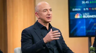 Jeff Bezos, a bald man in a dark suit and open-collared dark shirt, speaks and gestures with his right hand. He is seated against a warm, wood-paneled background, with a blurred financial news monitor showing 'NBC Business' visible behind him to the right.