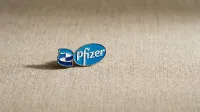 A blue and white Pfizer logo pin lies on a light brown, subtly textured fabric surface, casting a soft shadow. The oval pin features the word 'Pfizer' in white script on a blue field, with an abstract 'P' symbol in blue and white on the left.
