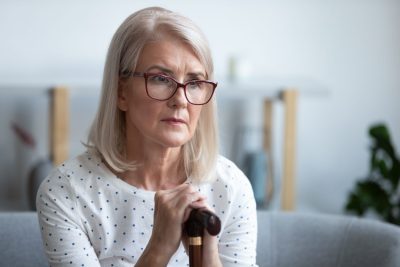 Sad lonely thoughtful pensive old mature woman hold cane stick sit alone on sofa, upset poor depressed senior retired grandma having health problem think of loneliness disease in retirement house