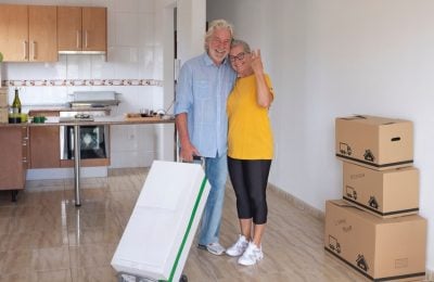 Smiling senior couple with white hair hugging gesturing ok sign in the new empty apartment with moving boxes on the floor - concept of active elderly people and new beginning like retired