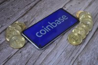 A smartphone displaying the white 'coinbase' logo on a blue screen lies diagonally on a light wooden surface. Several gold-colored Bitcoin coins are scattered to the left and right of the phone.