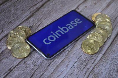 A smartphone displaying the white 'coinbase' logo on a blue screen lies diagonally on a light wooden surface. Several gold-colored Bitcoin coins are scattered to the left and right of the phone.