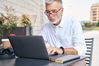 Focused older business man using computer working online outdoor. 60s serious businessman professional looking at laptop checking web market, elearning webinar, having remote hybrid call sits outside.