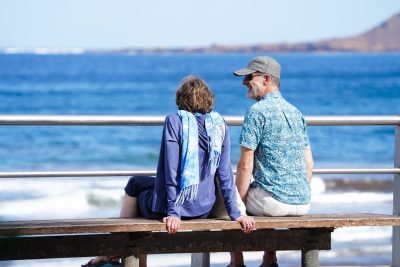 Couple of old retired seniors man and woman siting at a bench watching ocean by the beach and laughind or smiling