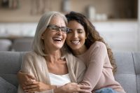 Happy excited senior mom and adult daughter woman having fun at home, hugging with heads touch, sitting on sofa, laughing with closed eyes, enjoying funny talk, leisure, family affection