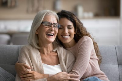 Happy excited senior mom and adult daughter woman having fun at home, hugging with heads touch, sitting on sofa, laughing with closed eyes, enjoying funny talk, leisure, family affection