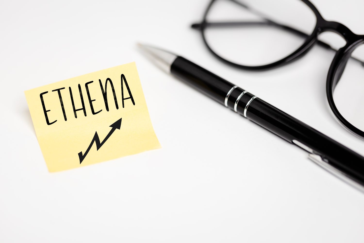 A yellow piece of paper on a white background with a handwritten inscription "ETHENA" and a growth chart, next to it a black pen and glasses (selective focus)