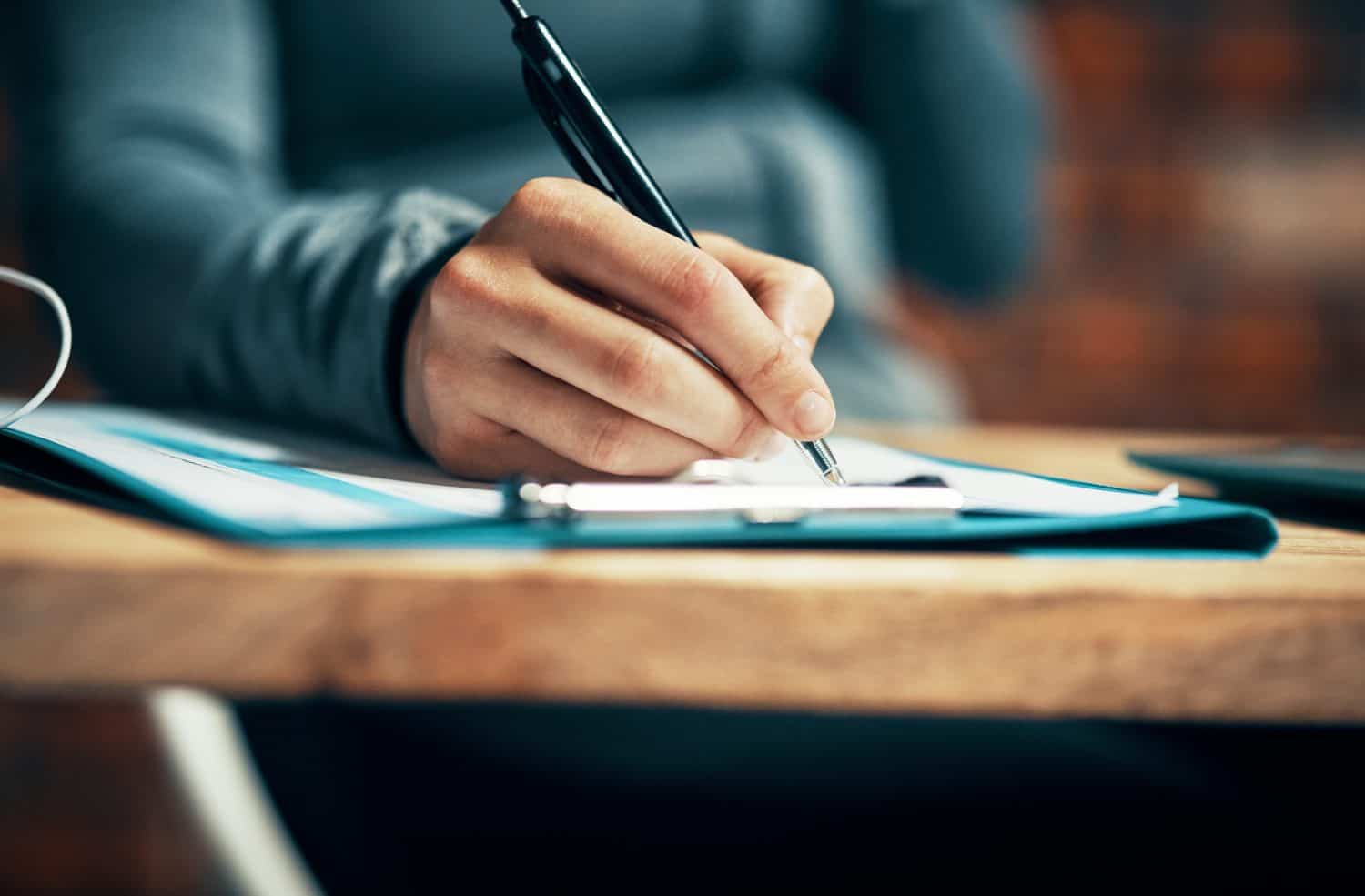 Hand, person and writing on clipboard on desk with filling application form or employment contact. Employee, tablet and pen with document or paperwork in file for signature for agreement in closeup