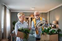 Senior couple carefully unpacking houseplant and wall clock from cardboard boxes, settling into new apartment and reminiscing about past memories together
