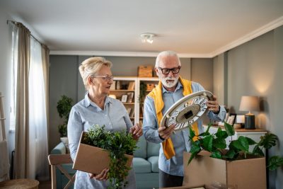 Senior couple carefully unpacking houseplant and wall clock from cardboard boxes, settling into new apartment and reminiscing about past memories together