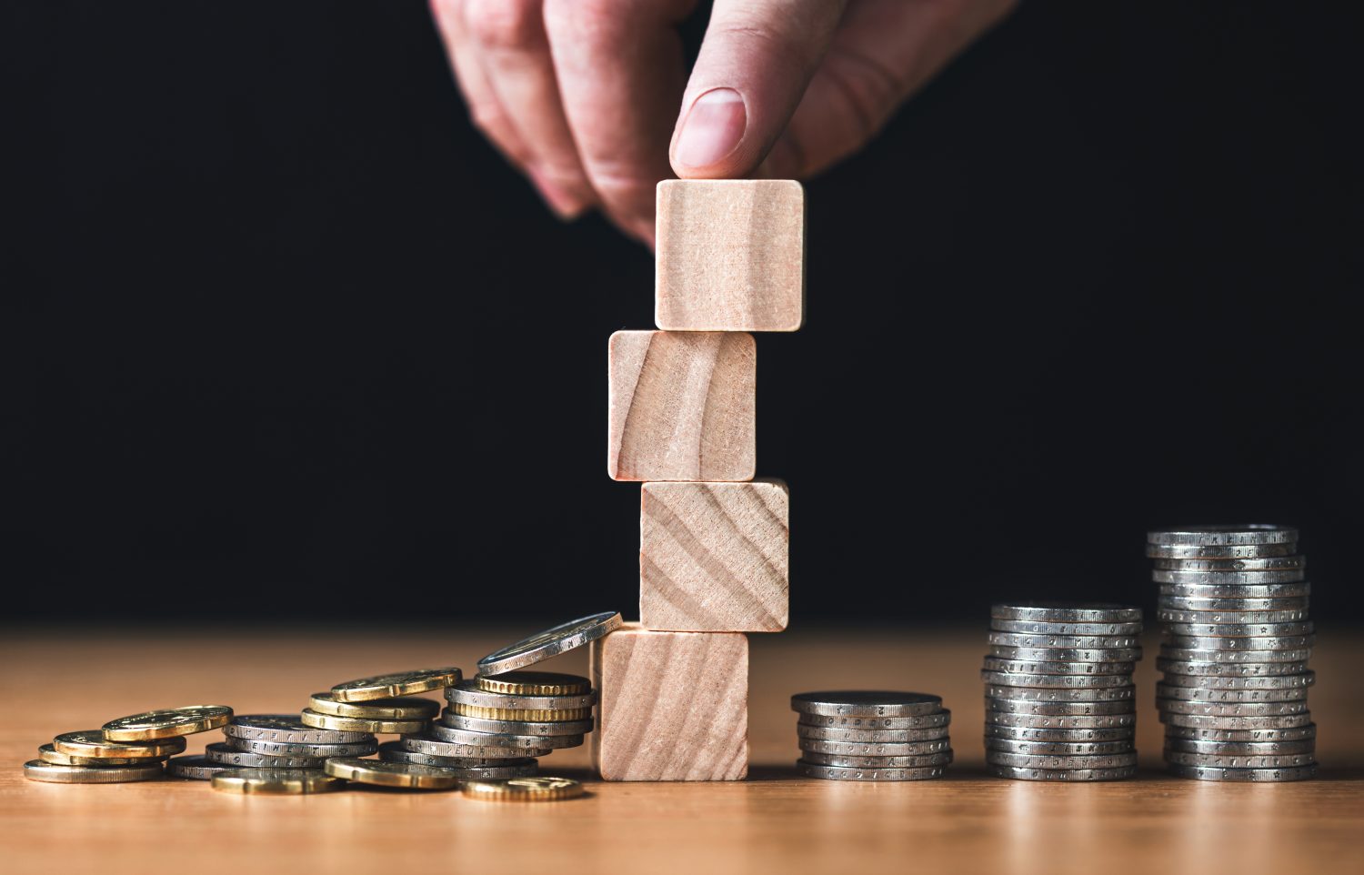 Hand stacks wooden blocks near coins. left money scattered, right coin stacks rising. Investment risk, volatility, wealth growth, portfolio strategy and risk management concept