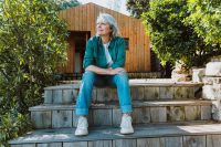 Senior woman relaxing on rustic wooden stairs, looking thoughtfully into the distance, embracing a peaceful outdoors retirement lifestyle at her simple wooden cabin surrounded by nature