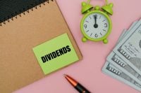 A green sticky note with text DIVIDENDS on a notebook, featuring a stack of US dollars, a pen, and an alarm against coloured background. Concept of passive income and time value of money