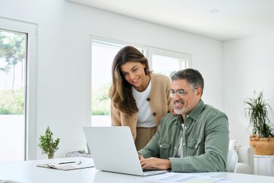 Smiling middle aged senior man working on computer sitting at table with wife standing nearby in living room. Happy mature older couple using laptop technology at home. Authentic candid shot.