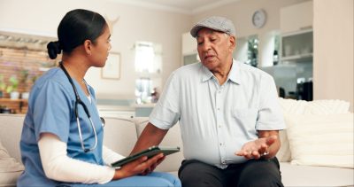 Elderly care, doctor and retired man on sofa for discussion, routine check up or diagnosis in living room. Female carer, senior patient and consultation on couch for healthcare, wellbeing or medicare