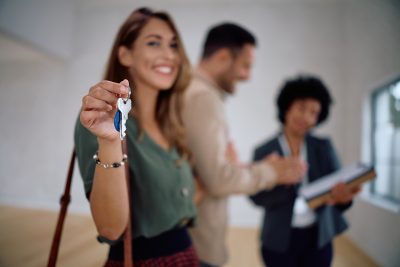 Close up of woman with new house keys. Her husband and real estate agent are in the background.