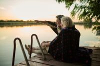 Retired couple sitting on a wooden pier by a lake. Covered with a cozy blanket. Savoring a peaceful sunset. Exuding love and romance in nature. Feeling tranquility outdoors. Relationship goals. Calm