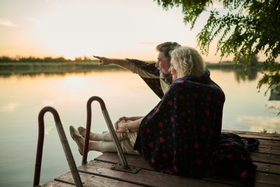 Retired couple sitting on a wooden pier by a lake. Covered with a cozy blanket. Savoring a peaceful sunset. Exuding love and romance in nature. Feeling tranquility outdoors. Relationship goals. Calm