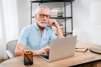 Serious older mature middle aged man wearing eyeglasses looking at computer technology sitting at table, using laptop hybrid working online, elearning, browsing web, searching online at home.