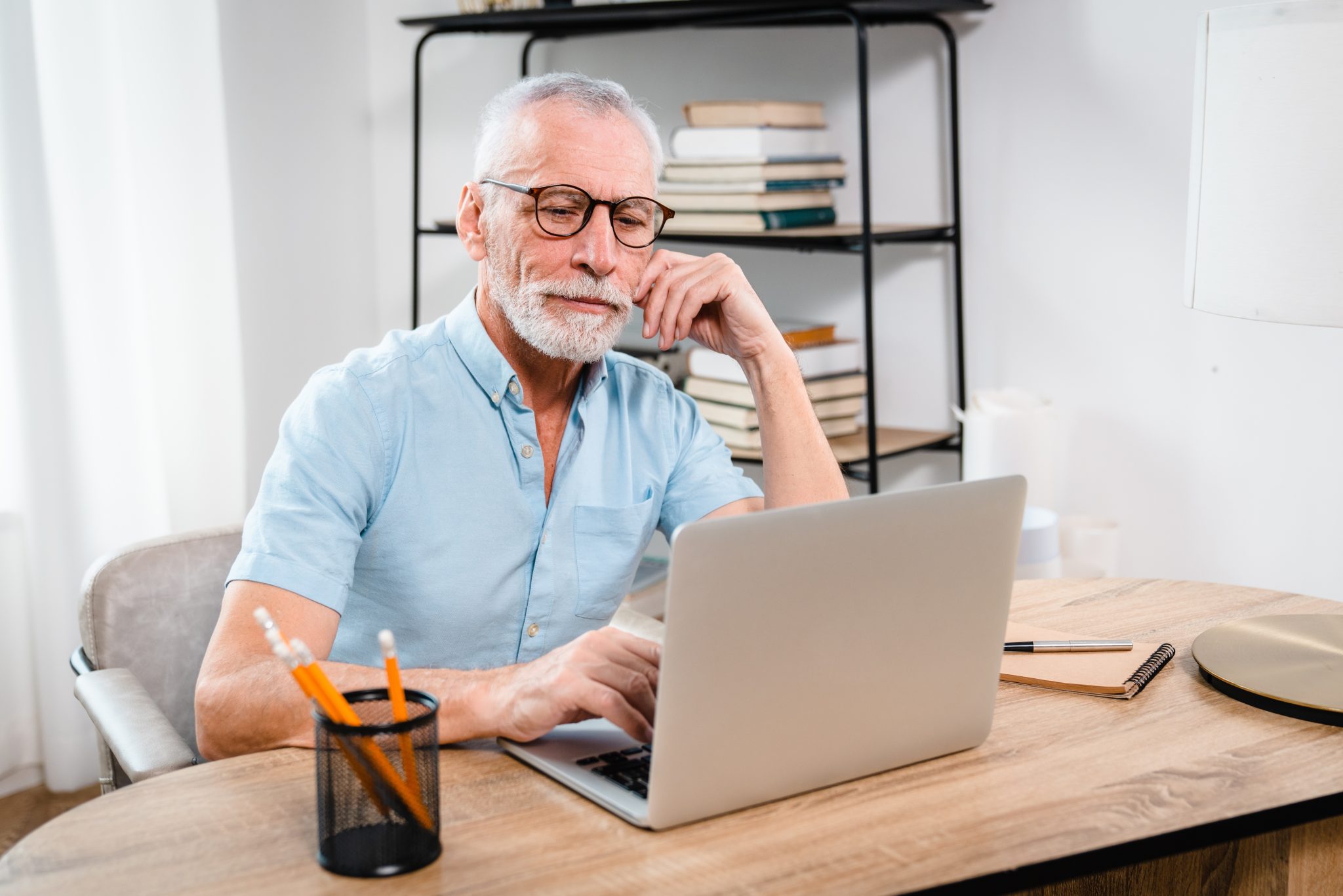 Serious older mature middle aged man wearing eyeglasses looking at computer technology sitting at table, using laptop hybrid working online, elearning, browsing