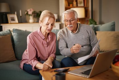 An elderly couple, a man and woman, are sitting on a sofa in their living room. The man is holding paperwork and a pen, pointing at something on the page.
