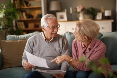 A mature husband and wife are seated on a couch, holding documents. The wife points at a paper while discussing finances with her husband in the comfort of their home living room.