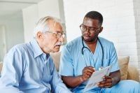 Doctor or nurse caregiver talking to senior man and showing a document or a brochure at home or nursing home