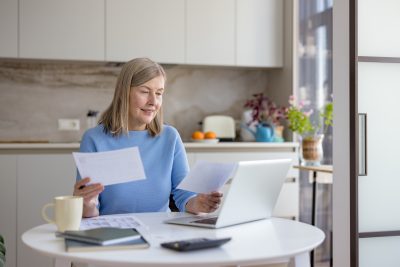 Senior woman at home managing finances, reviewing bills and documents at a desk with laptop and calculator, budgeting, planning retirement and household expenses with calm focus
