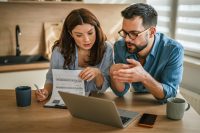 Young couple managing household finances, reviewing documents, and using a laptop to plan expenses and investments while working together in their modern kitchen